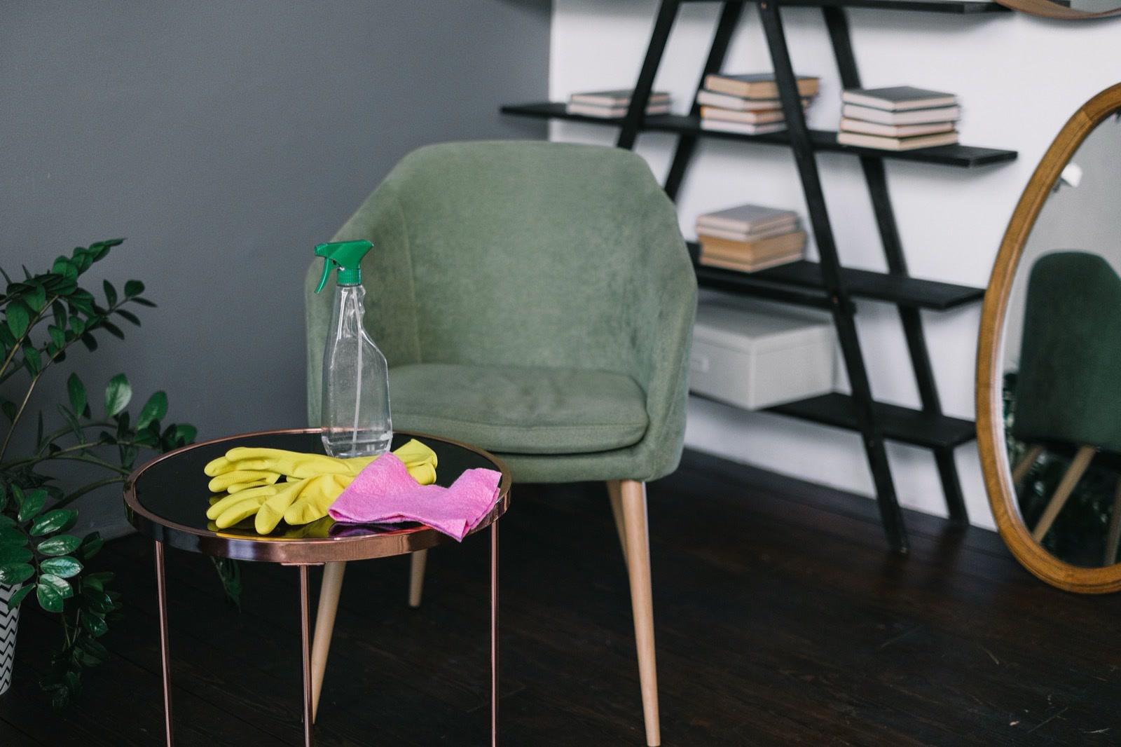 Yellow rubber gloves, a pink cloth and a spray bottle on a small side table beside a green armchair in a living room