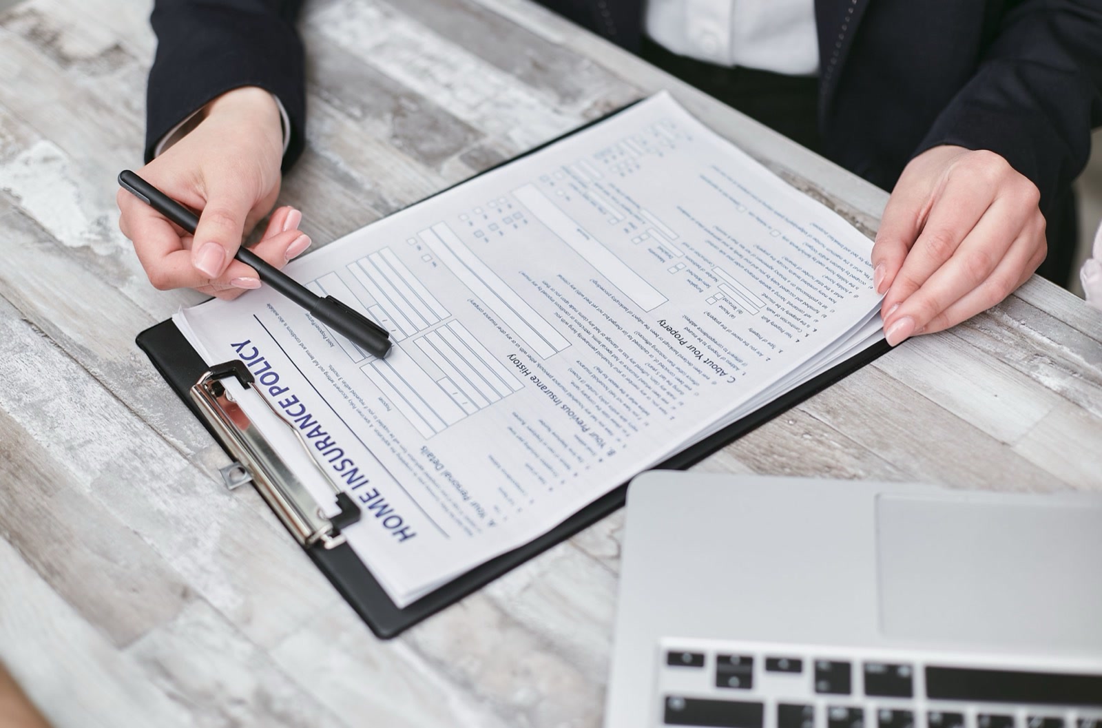 Hands holding a pen over an insurance policy form on a clipboard, beside a laptop on a wooden desk