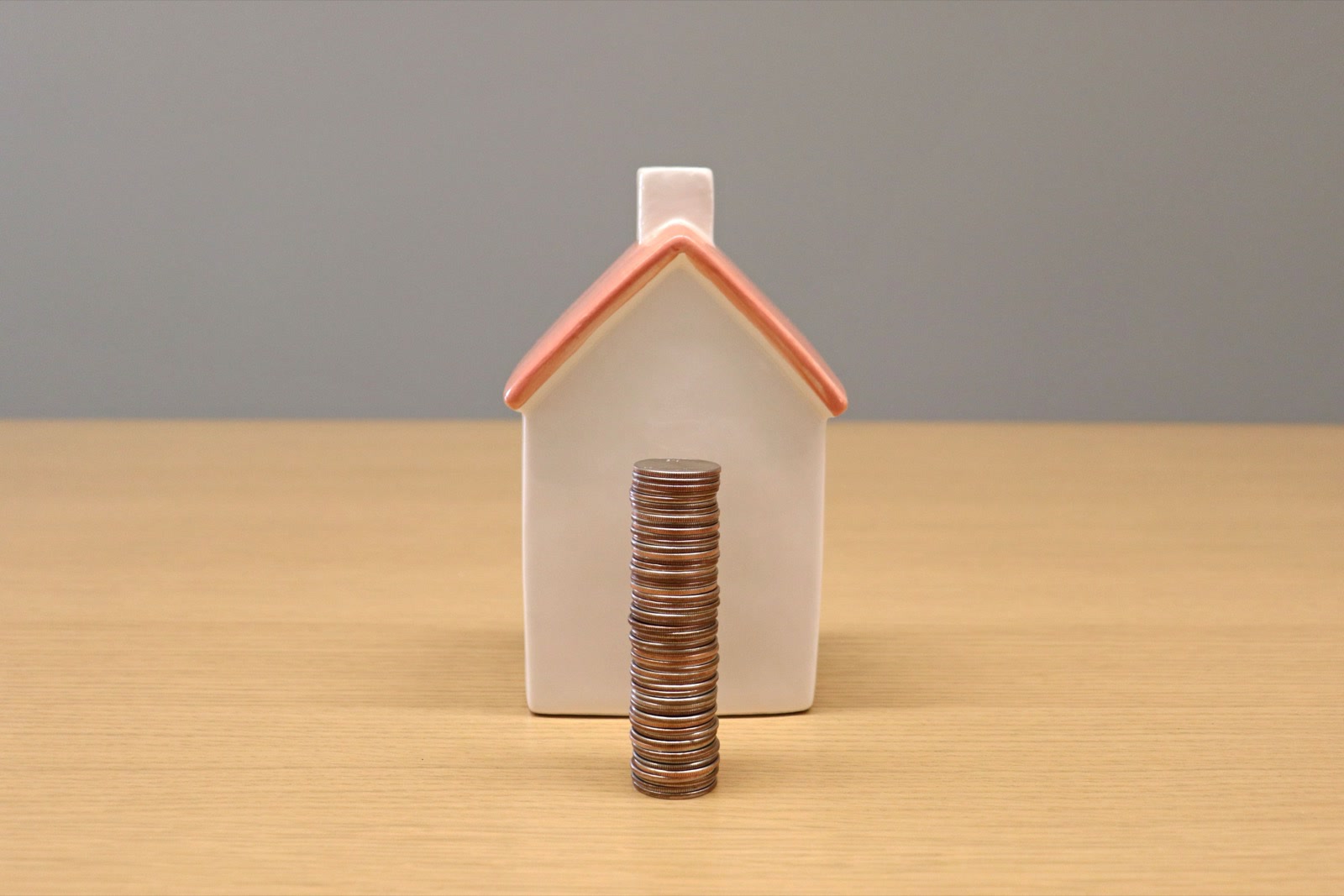 A small white ceramic house with a red roof behind a tall stack of coins on a wooden table