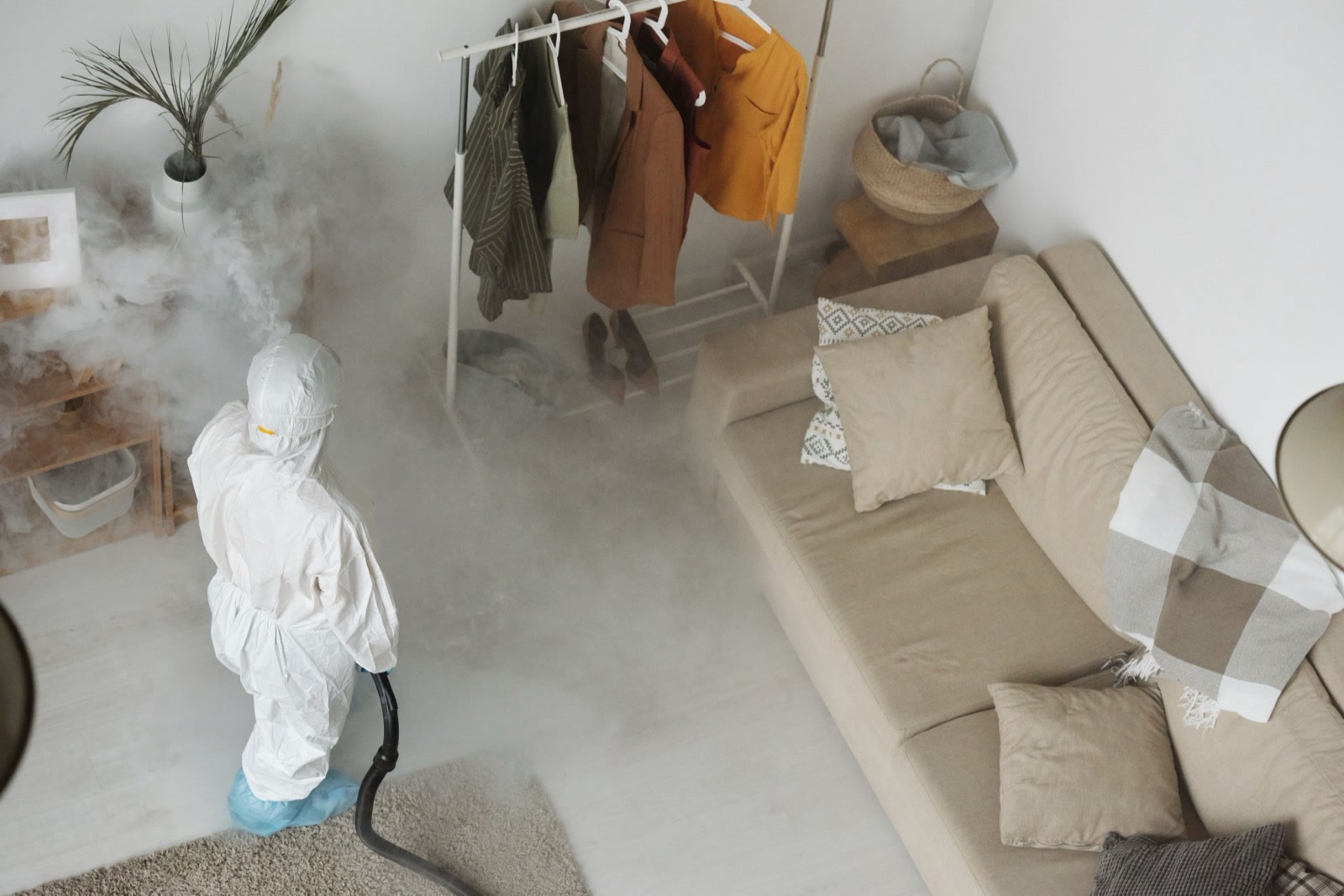A person in a white protective suit fogging a living room, seen from above, beside a sofa and a clothes rail