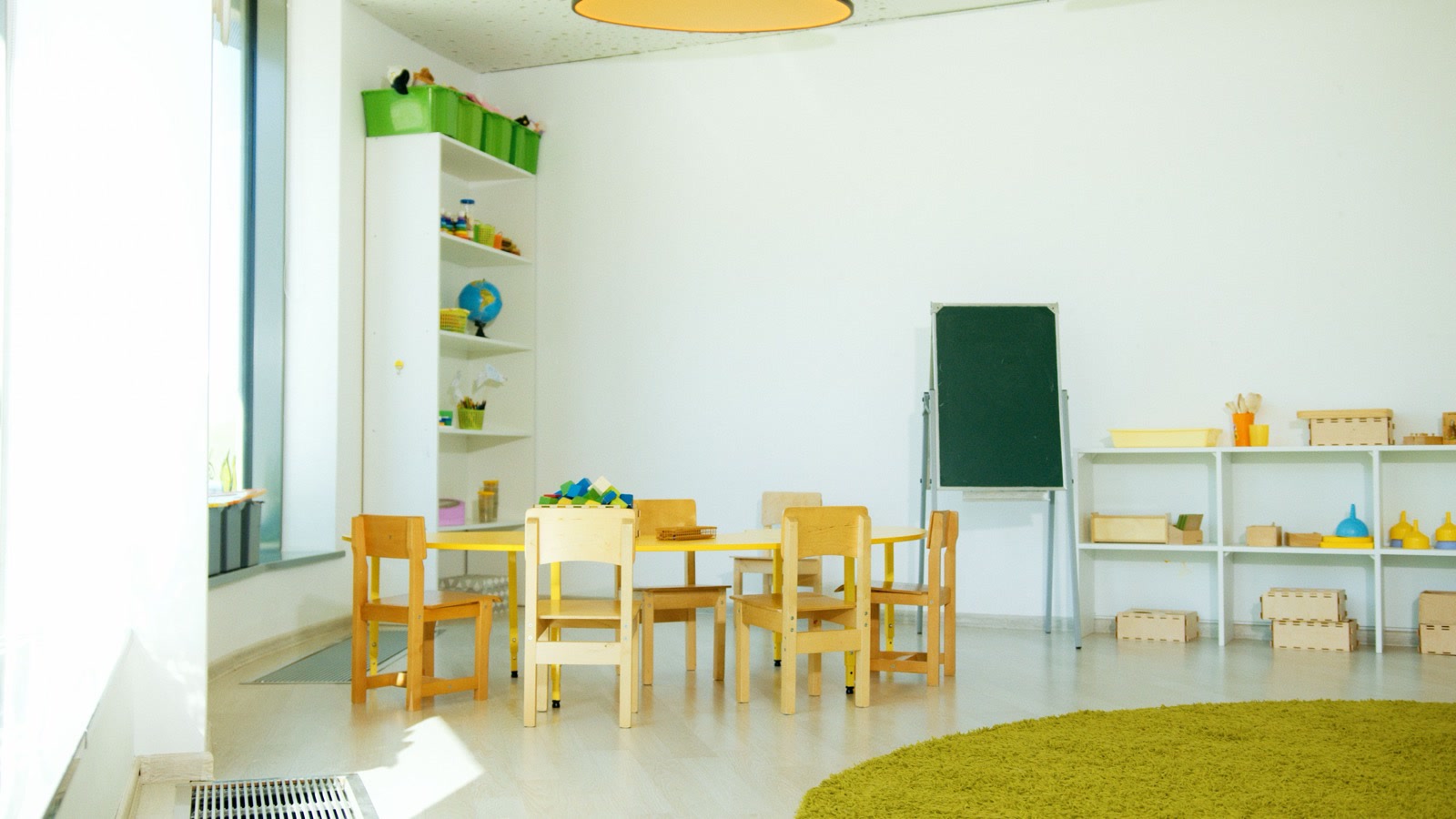 An empty, bright preschool classroom with small wooden chairs round a yellow table, a chalkboard and shelves of toys