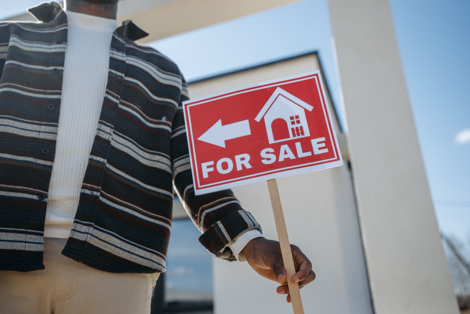 A person, seen from the chest down, holding a red For Sale sign in front of a house