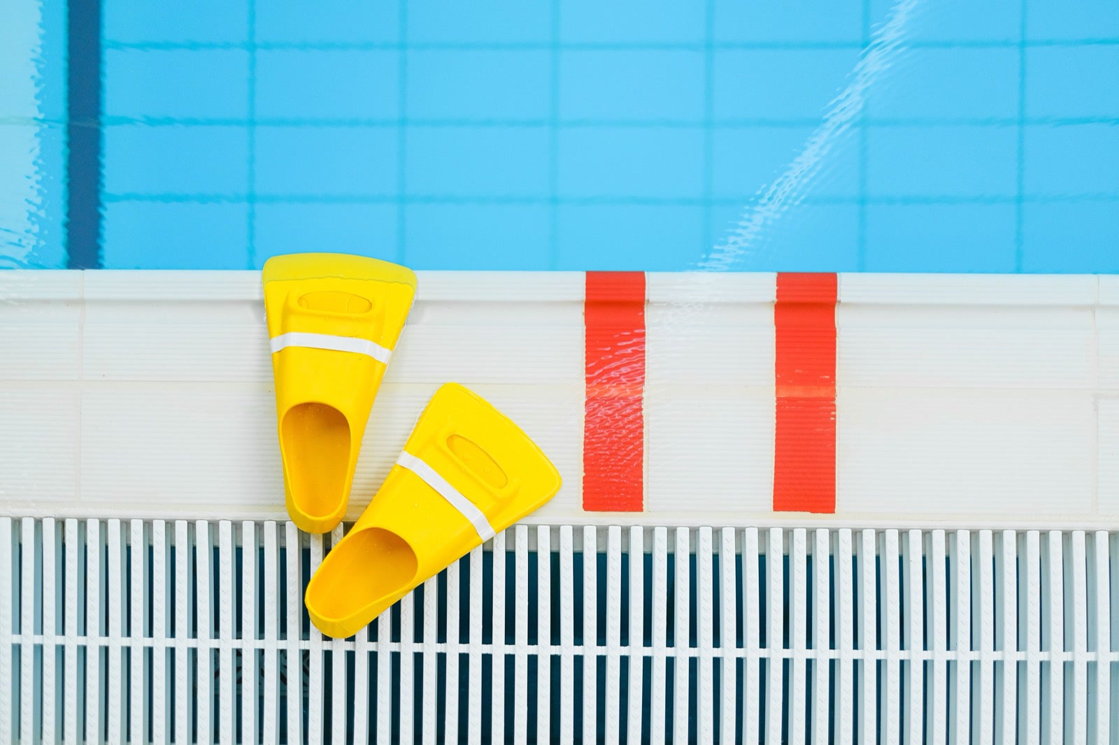 A pair of small yellow swim fins on the edge of an indoor pool, beside red lane markings